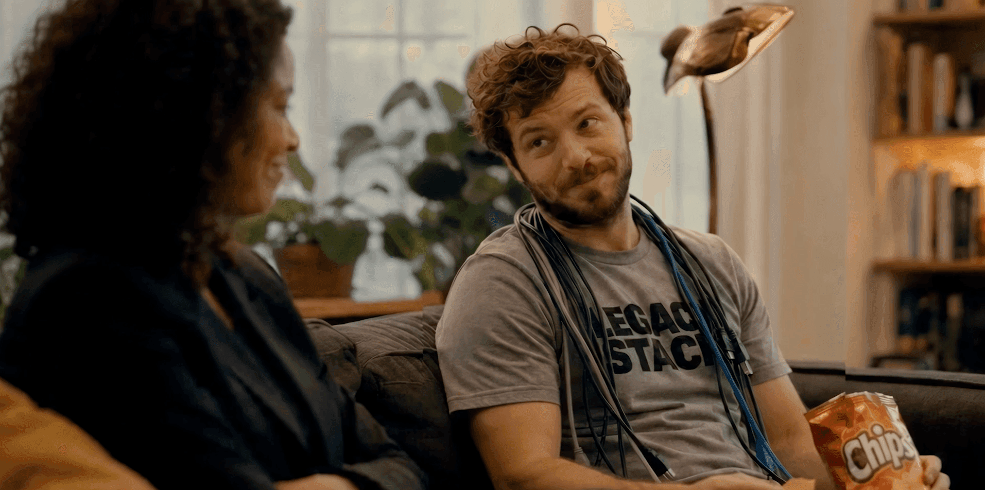 A man on a couch, holding a bag of chips, smiles and looks at a woman.