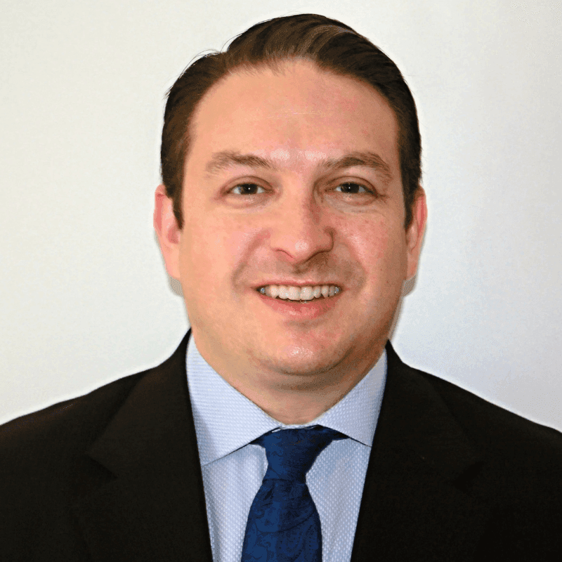 Smiling man in a dark suit and patterned tie, looking directly at the camera.