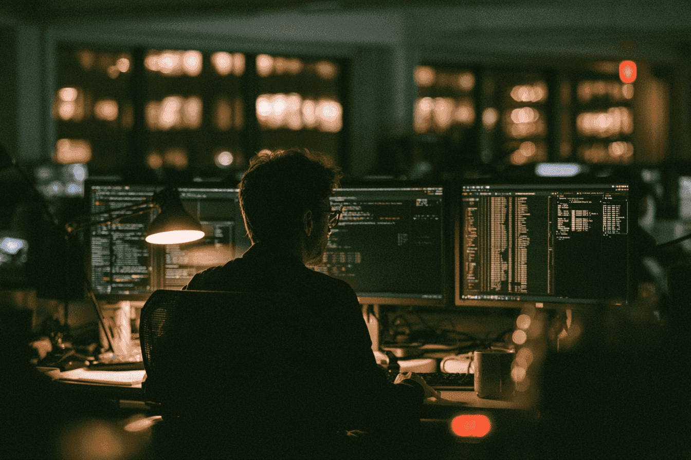 Silhouetted person working at dual computer monitors in a dark office setting.