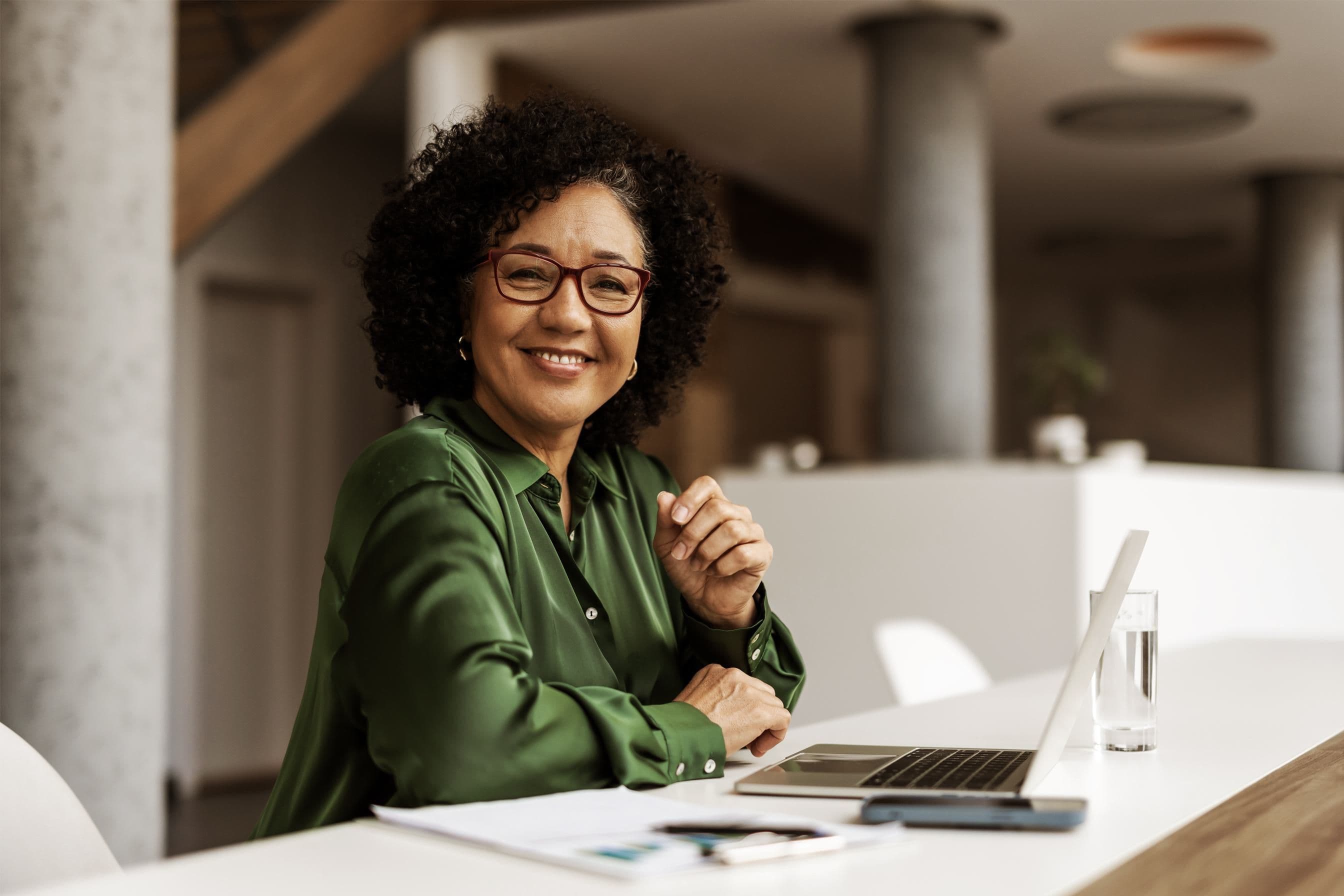 Smiling woman with curly hair and glasses works on a laptop in a modern office.
